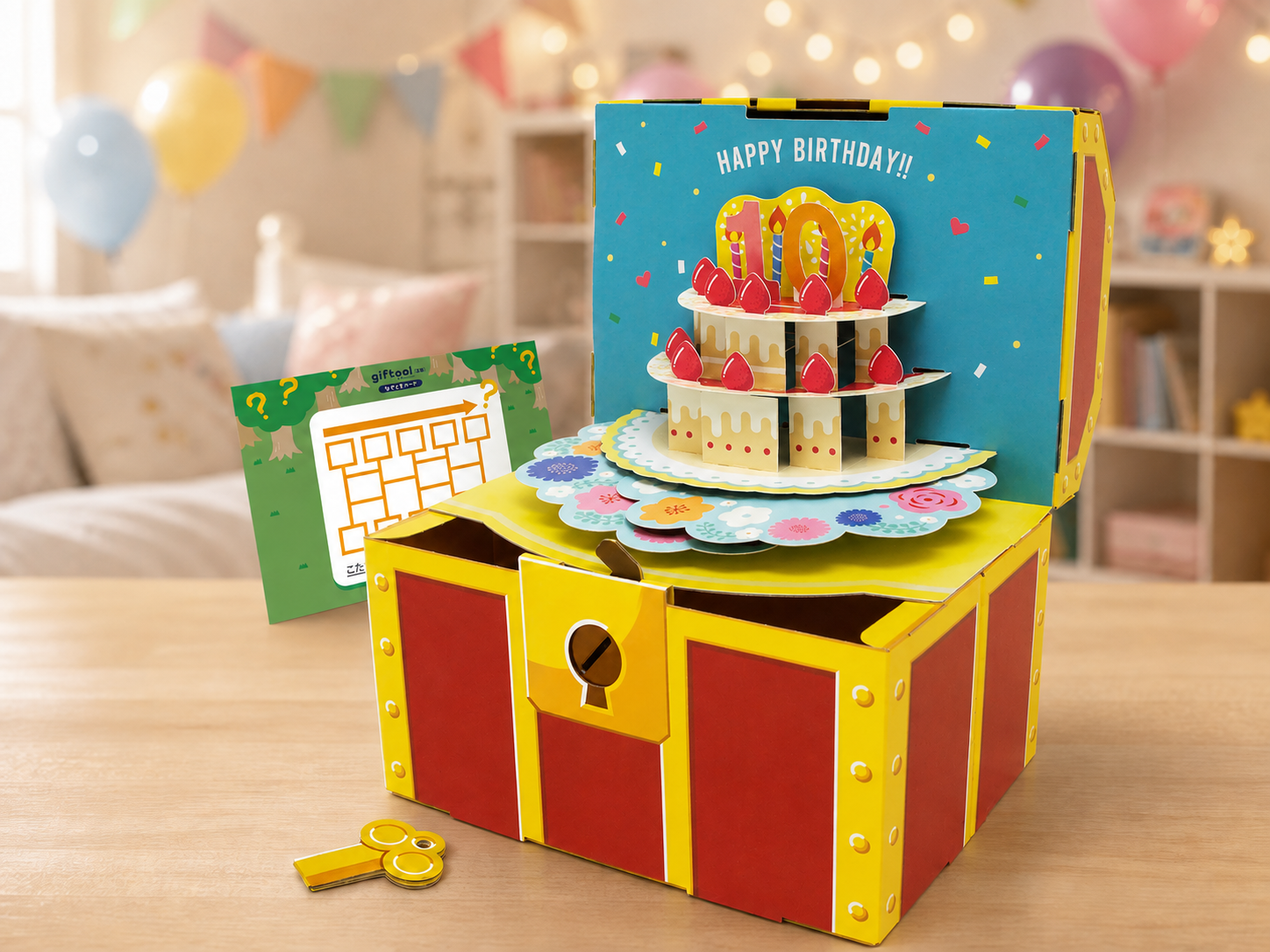 誕生日の飾り付けがされた子供部屋を背景に、ケーキが飛び出す宝箱となぞときシート、鍵を写した商品イメージ